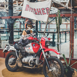 Dead Dakar Doug sitting on a Dakar BMW R100 holding a Bike Shed flag